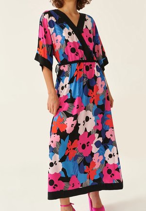 Woman wearing a colorful floral midi dress with black trim and pink high-heeled sandals, standing against a plain background.