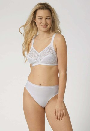 Donna con capelli biondi lunghi che indossa un reggiseno in pizzo bianco e mutandine bianche a vita alta abbinate, sorridente con le mani rilassate lungo i fianchi.