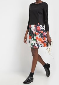 Svart långärmad topp i kombination med en blommönstrad kjol i livfulla färger. Slät tygtextur och eleganta svarta skor med spännen.