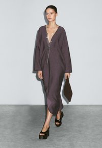 Lila Leinenkleid mit tiefem V-Ausschnitt, langen Ärmeln und seitlichen Schlitzen. Das Modell trägt eine braune Clutch und schwarze Plateauschuhe.