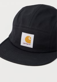 Μαύρο καπέλο από ανθεκτικό ύφασμα με επίπεδη κυρτή, σχεδιασμό με έξι πάνελ και λευκή τετράγωνη κονκάρδα με κίτρινο λογότυπο και κείμενο.