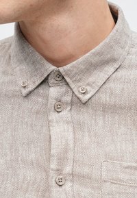 Košeľa so vzorom rybej kosti v svetlo béžovej látke, s golierom na gombíky a štyrmi béžovými gombíkmi pozdĺž zapínania.