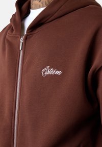 Brauner Reißverschluss-Hoodie aus weichem Stoff, mit einem weißen, gestickten Logo im linken Brustbereich und einem standardmäßigen Kapuzen-Design.