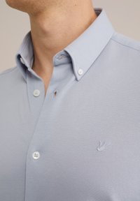 Světle modrá košile s límečkem z texturované látky, vybavená bílými knoflíky a malým vyšitým logem na hrudi.