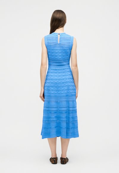 Femme debout de dos, portant une robe midi bleue sans manches à motif avec un détail clé à l'arrière et des ballerines imprimé léopard.