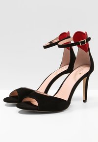Czarne zamszowe sandały na wysokim obcasie z otwartymi palcami, z paskami na kostkach i złotymi klamrami oraz czerwonymi serduszkowymi akcentami na obcasach.