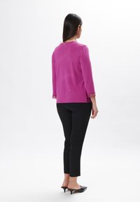 Fuchsia Pullover mit dreiviertel Ärmeln, filigranen Spitzenverzierungen an den Bündchen und einem kleinen goldenen Knopf hinten. Kombiniert mit schwarzen Slim-Fit-Hosen.