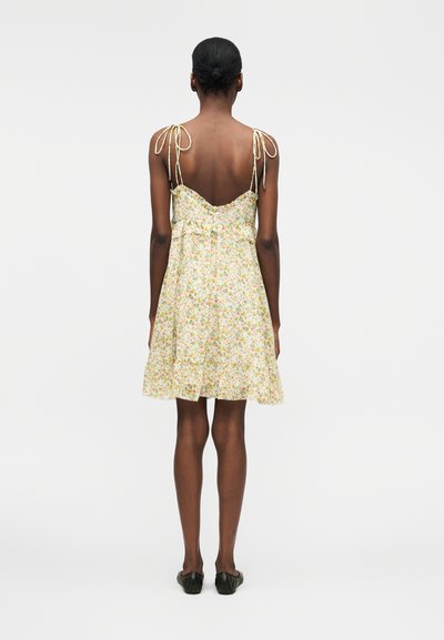 Γυναίκα με σκουρόχρωμο δέρμα και μαλλιά σε κότσο φοράει ένα ανοιχτό floral φόρεμα με λεπτές ράντες, χαμηλή πλάτη και βολάν στο τελείωμα, στέκεται μπροστά σε απλό φόντο.