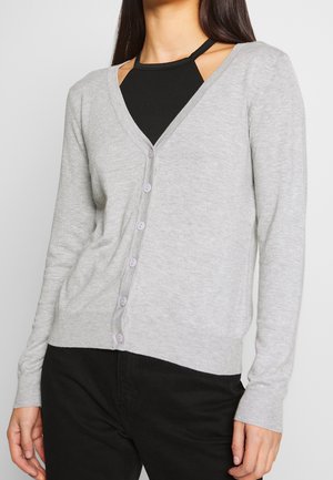 Femme portant un cardigan gris clair boutonné par-dessus un haut noir à col halter, associé à un pantalon noir, debout sur un fond uni.