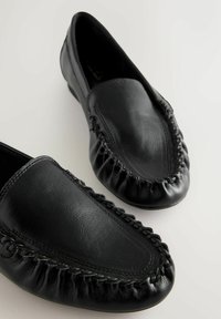 Mustad nahast mokassiinid ümarate varvastega, millel on õmmeldud äär ülaosas ja sile tekstuur. Tasase tallaga, stiilse disainiga.