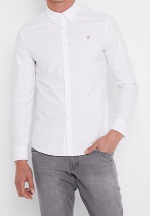 Homme portant une chemise blanche ajustée avec petit col et un jean gris, main dans la poche, sur un fond blanc uni.