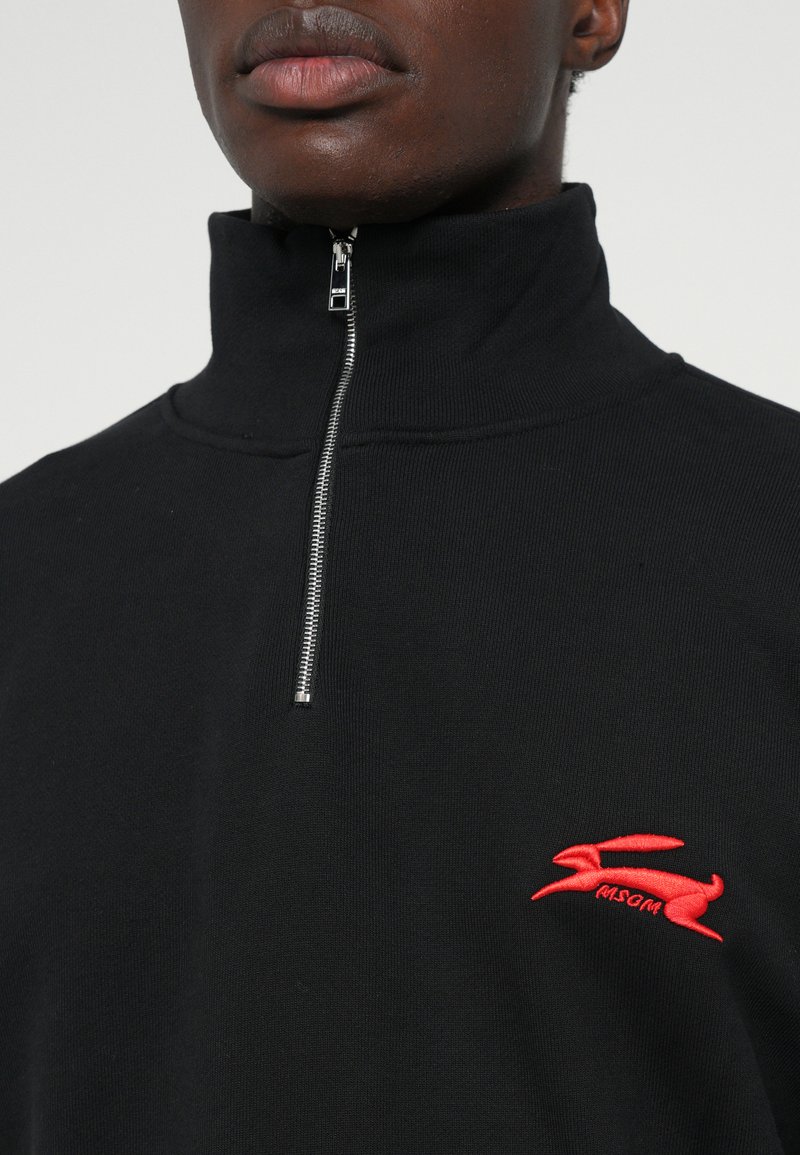 Man in een zwart zip-up vest met hoge kraag en een rood geborduurd konijnenlogo op de borst.