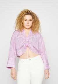 Blusa cropped em roxo claro com padrão geométrico, apresentando um fecho de cordão na frente, mangas largas e textura suave. Combinada com calças brancas.