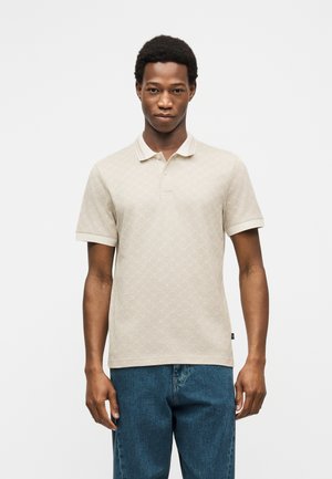 Jeune homme portant un polo beige à motifs et un jean bleu, debout devant un fond blanc uni.