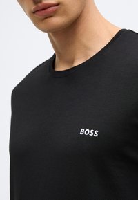 Μαύρο βαμβακερό μπλουζάκι με στρογγυλή λαιμόκοψη και λευκό κεντημένο λογότυπο «BOSS» στην αριστερή πλευρά του στήθους. Λείο, εφαρμοστό σχέδιο.