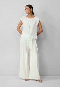 Witte laagjesblouse met korte ruches mouwen, gecombineerd met ruime witte broek en sandalen met bandjes, tegen een grijze achtergrond.