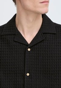 Cămașă scurtă cu mâneci scurte, de culoare neagră, cu un material texturat și model waffled, guler ascuțit și două nasturi din lemn pe plastronul din față.