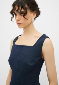 Dunkelblaues Denimkleid mit quadratischem Ausschnitt, strukturierten Trägern und genähten Details. Das Model trägt silberne Creolen.