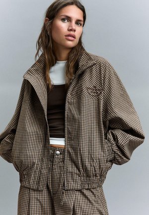 Tânără purtând un costum maro și bej în carouri, format din sacou și pantaloni, peste mai multe straturi de bluze albe și maro, stând în fața unui fundal simplu, gri.