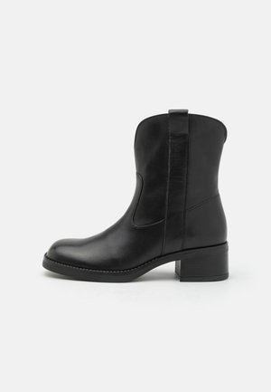 Botte cheville en cuir noir avec un bout arrondi, des panneaux latéraux élastiques et un petit talon carré. Texture lisse avec des coutures minimalistes.