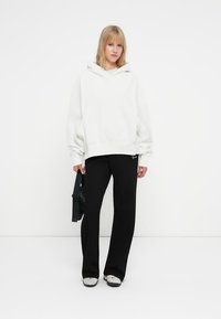 Felpa oversize bianca con tasca frontale, abbinata a pantaloni larghi neri e sneakers grigie. Il modello tiene una borsa nera. Design minimalista.