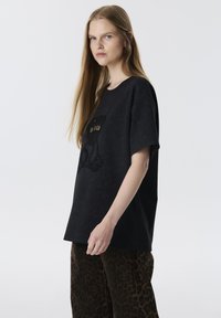 Zwarte t-shirt met een beerafbeelding en de tekst "BOLD", met korte mouwen en een losse pasvorm. Gepaard met een broek met een bruin luipaardprint.