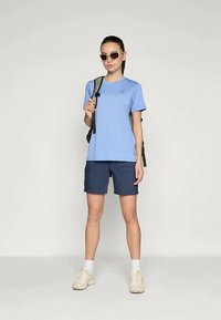 Kvinna som bär en ljusblå t-shirt, marinblå shorts, vita sneakers, solglasögon och bär en ryggsäcksrem över ena axeln, stående mot en enkel bakgrund.