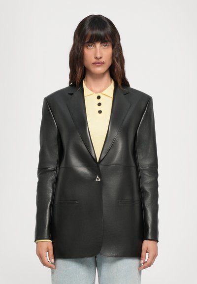Blazer de cuero negro con un diseño estructurado, que presenta un sutil broche triangular, bolsillos frontales y una textura suave. Llevado sobre un polo amarillo claro.