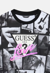 Black and white graphic T-shirt featuring a triangular "GUESS" logo. Text "LOVE" in pink script; cotton material with black neckline.