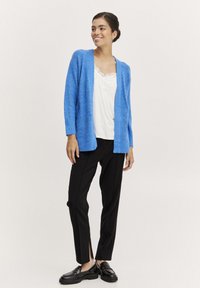 Blå, fluffig cardigan med öppen front, parat med en vit blus med spetsdetaljer och svarta skräddarsydda byxor med sidslitsar, komplett med svarta loafers.