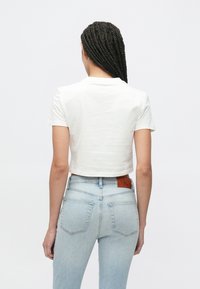 Fehér, rövid ujjú, rövid fazonú póló, világoskék magasderekú farmernadrággal párosítva, amelyen hátul zsebek és barna bőrcímke található.