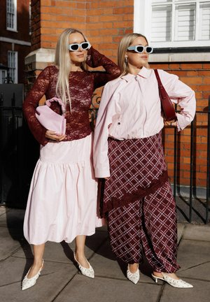 Două femei purtând ochelari de soare și ținute cu modele în burgund și roz, stând pe un trotuar cu clădiri din cărămidă în fundal.
