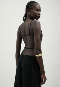Top in tessuto trasparente nero con spalline sottili, abbinato a pantaloni neri larghi. Un notevole bracciale a manica dorato mette in risalto il braccio.