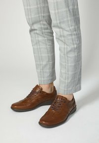 Brązowe skórzane trampki z zaokrąglonym noskiem, płaskimi sznurowadłami i przeszyciami. W zestawie z jasnoszarymi spodniami w kratę.