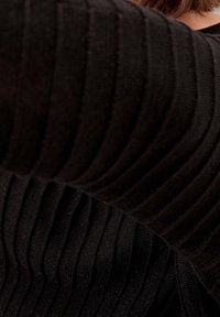 Gros plan d'une personne portant un pull noir côtelé, le bras levé, couvrant partiellement son cou et ses cheveux.