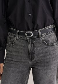 Črn usnjen pas z ovalno srebrno sponko, nošen preko sivih kavbojk z visokim pasom in rahlo zbledelo teksturo ter petimi žepi.