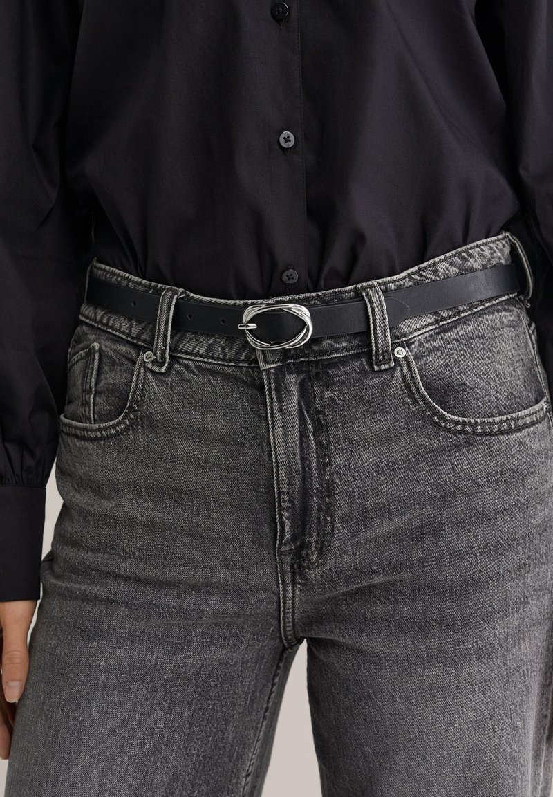 Črn usnjen pas z ovalno srebrno sponko, nošen preko sivih kavbojk z visokim pasom in rahlo zbledelo teksturo ter petimi žepi.