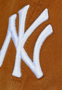 La tela de algodón marrón presenta un logo bordado en blanco con la inscripción "NY". La textura es acanalada con detalles de costura visibles.