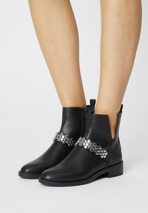 Bottines noires en cuir avec un petit talon et un détail de chaîne hexagonale argentée sur le devant, portées par une personne avec des jambes nues sur fond blanc.