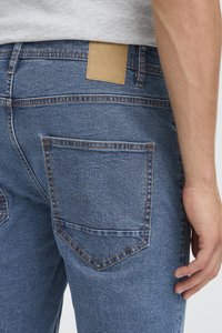 Detailní záběr na zadní kapsu a pas džínsů s hnědou koženou záplatou, nošené osobou s uvolněnou rukou u boků.