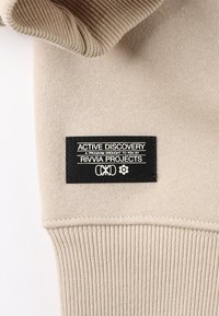 Article d'habillement beige avec une texture côtelée. Présente une étiquette noire portant l'inscription "ACTIVE DISCOVERY" en texte et graphiques blancs.