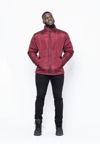 Giacca piumino rossa con colletto alto, zip frontale e tasche laterali; indossata sopra jeans neri e stivali neri; texture lucida e dettagli di cucitura.