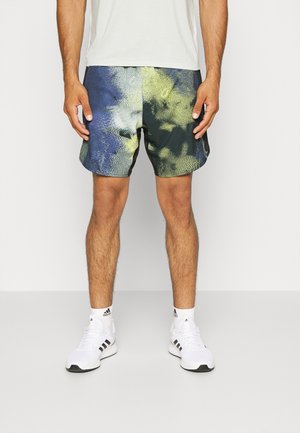 Männliche Person trägt mehrfarbige blaue und gelbe Sportshorts, weiße Adidas-Sneaker und weiße Socken, steht vor einem schlichten hellen Hintergrund.