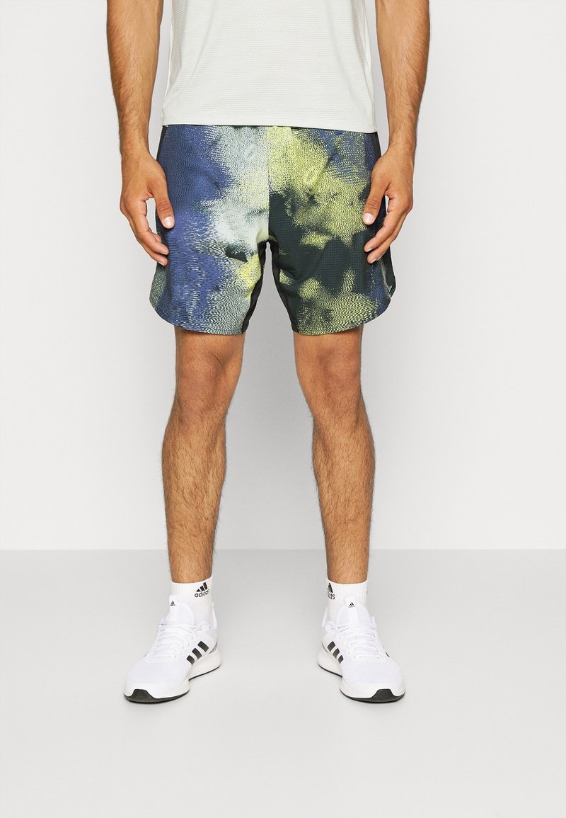 Homme portant un short de sport multicolore bleu et jaune, des baskets blanches Adidas et des chaussettes blanches, debout devant un fond clair uni.