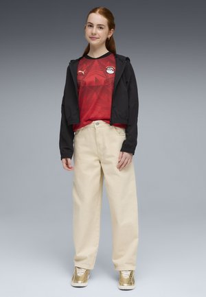 Punahiuksinen nuori, joka pukeutunut mustaan takkiin punaisen Puma-urheilupaidan päällä, beigen väriset leveälahkeiset housut ja kultaiset lenkkarit, seisoo harmaan taustan edessä.