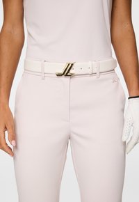 Donna con pantaloni rosa pallido e top abbinato, cintola bianca con fibbia dorata JL e guanto in pelle bianca perforata.