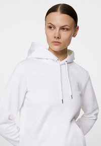 Vit luvtröja med en framsida ficka, dragsko med metallspetsar och en diskret logotypdetalj på bröstet. Slät tygtextur.