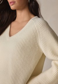 Svetlobéžový krémový sveter s výstrihom do V, ktorý má vertikálne rebrovanie, jemnú textúru a voľný strih, s viditeľnými švovými detailmi pozdĺž výstrihu a rukávov.