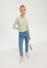 Light green long-sleeve top, fitted design. Light blue high-waisted jeans, cropped length. Gray sneakers with purple accents, holding a small gray bag.