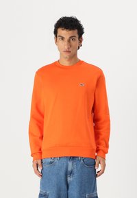 Hanorac portocaliu cu guler rotund, mâneci lungi, manșete ribbate și un mic logo verde pe piept. Confecționat dintr-un material neted și elastic.
